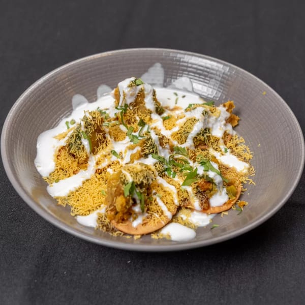 A grey bowl of Indian chaat, topped with white sauce, crispy noodles, and green herbs.