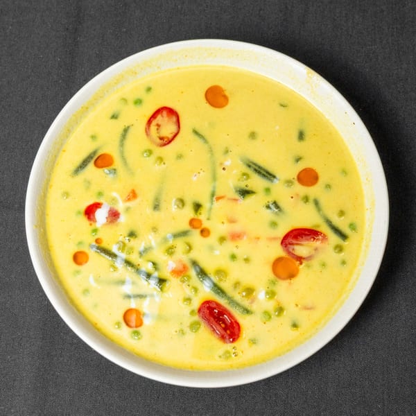 A white bowl filled with yellow curry, green peas, red tomatoes, and green beans.