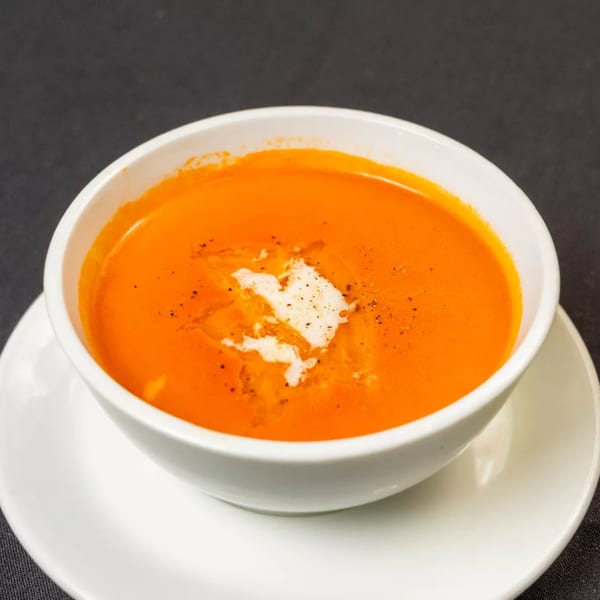 A white bowl of vibrant orange tomato soup, garnished with a swirl of cream and black pepper.