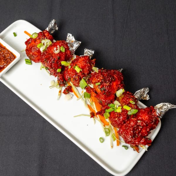 Six vibrant red chicken lollipops with foil-wrapped bones, garnished with green onions and shredded vegetables.