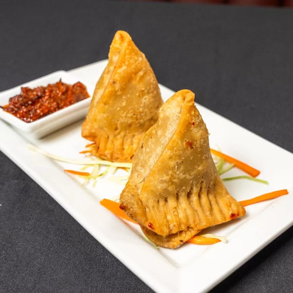 Two golden-brown samosas with a side of red chutney on a white rectangular plate.